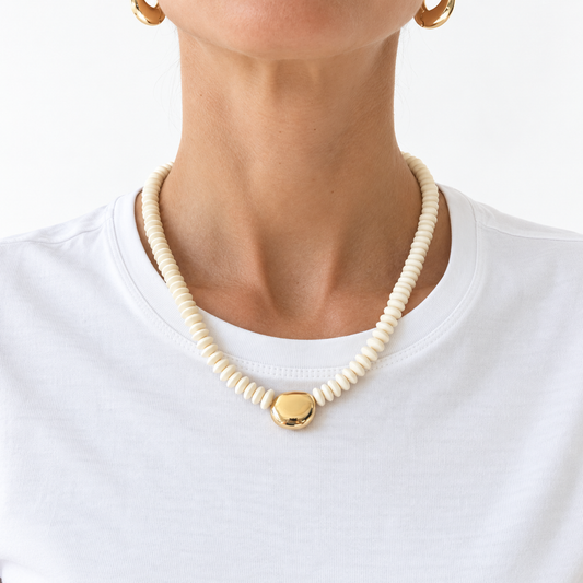 A close-up of a model wearing a cream-colored beaded necklace with a polished gold pendant, paired with a white t-shirt and gold hoop earrings.