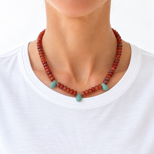 A close-up of a person's neck and upper chest wearing a beaded necklace. The necklace consists of small reddish-brown beads with three light green teardrop-shaped accent beads at the center front, worn over a white crew-neck shirt against a plain white background.