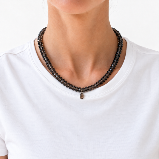 A close-up of a person wearing a double-strand beaded necklace made of dark brown to black faceted beads with a small oval pendant, paired with a white crew-neck t-shirt.