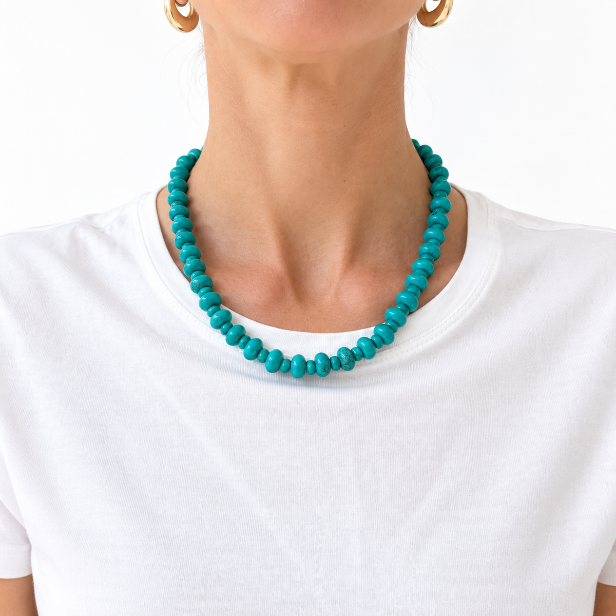 A close-up of a model wearing a vibrant turquoise beaded necklace with small, rounded disc-like beads, paired with a white t-shirt and gold hoop earrings.